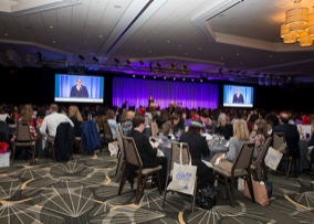 conference attendees sitting at tables watch a speaker projected on a large screen