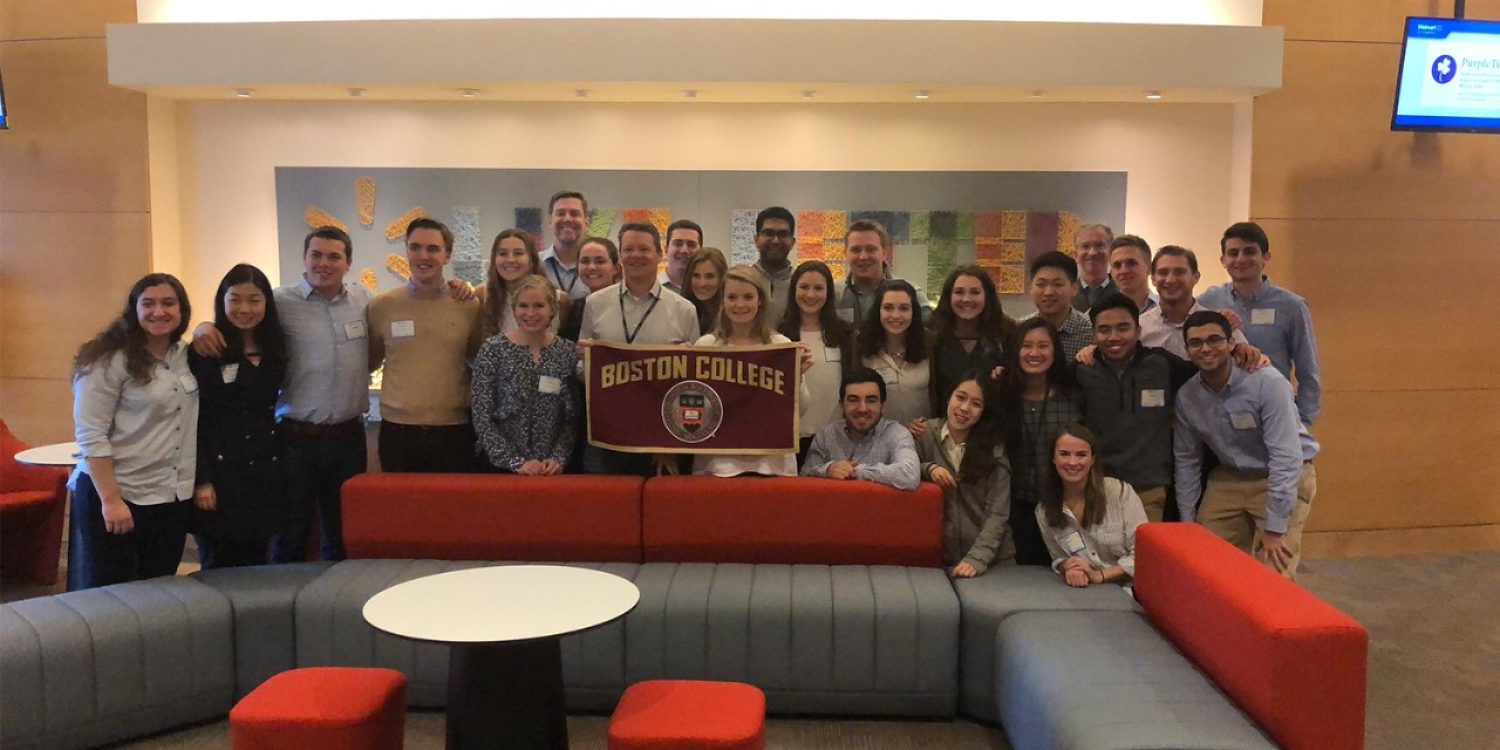 a group of students post in a company lobby while holding a Boston College sign