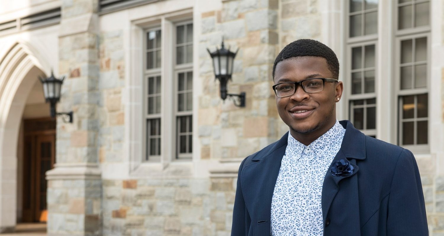 Kofi Nimo, a young black man in glasses and a button down with a jacket, stands in front of Fulton Hall