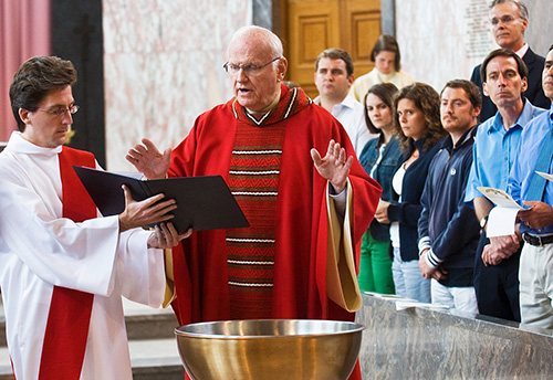 Priest in red with hands raised to bless a font, a deacon holding the text to read, with congregants to the right.