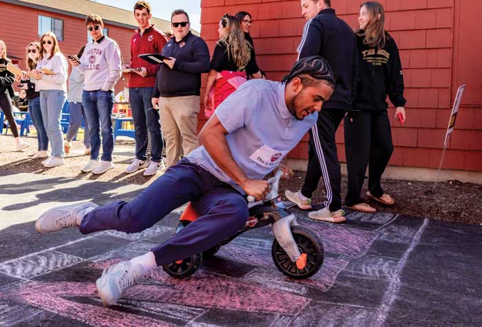 Miles Hester ’24 crosses a chalk drawn finish line on a trike with fellow student onlookers.